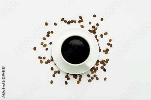 A white cup with a saucer and black coffee, coffee grains are scattered around on a white background. Flat lay, top view