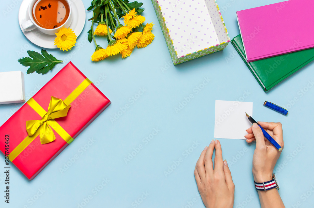 Flat lay top view womens hands write congratulations on empty sheet, red present box with gold ribbon, cup of tea, notebook, yellow chrysanthemums on blue background. Holiday concept, Congratulations Flat lay top view womens hands write congratulations on empty sheet, red present box with gold ribbon, cup of tea, notebook, yellow chrysanthemums on blue background. Holiday concept, Congratulations