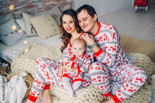 Parents with their little daughter in holiday clothing with printed deers and snowflakes sitting on the bed in cozy room with Christmas tree, gifts and Christmas lights. New year and Christmas concept