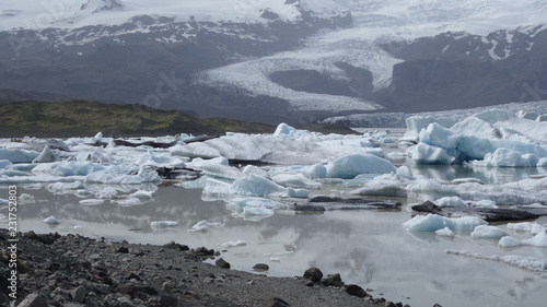 Eisberge auf einem Fluß in Island hinter schwarzem Felsufer und vor Gebirgslandschaft