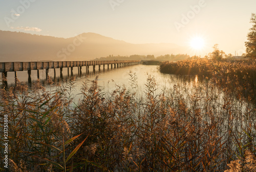 long wooden boardwalk pier over water in golden evening light with a mountain landscape silhouette in the background and golden marsh grass in the foreground