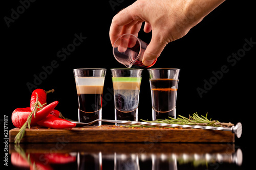 Tableau sur toile Three alcohol shooter layered cocktails on a wooden board served with barman spoon and chili pepper isolated at black background