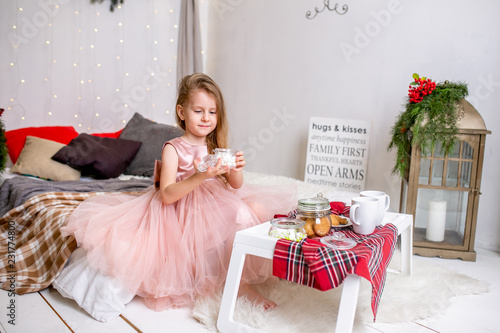 Pretty little girl 4 years old in a pink dress. Child in the Christmas room with a bed, eating candy, chocolate, cookies and drinking tea, feeding teddy bears and laughing, good mood