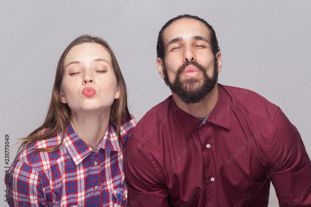 Portrait of funny bearded man with black collected hair and woman in casual style standing with closed eyes and kissing to camera. indoor studio shot, isolated on gray background. Portrait of funny bearded man with black collected hair and woman in casual style standing with closed eyes and kissing to camera. indoor studio shot, isolated on gray background.