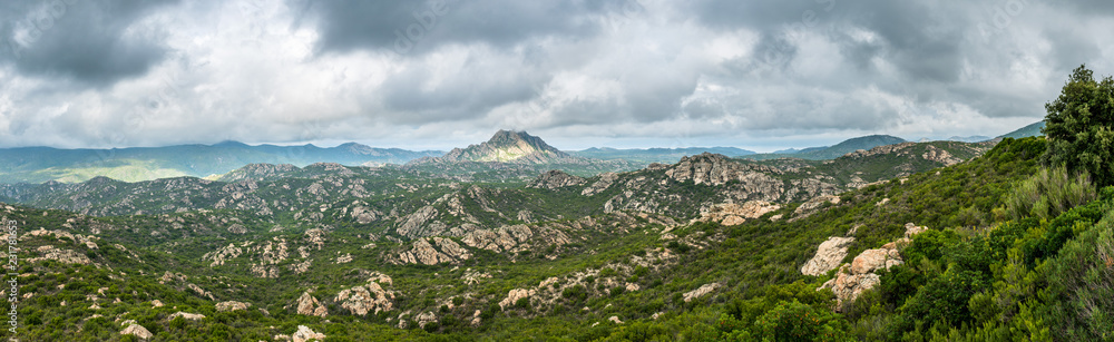 Panoramic view of Monte Genova in the Desert des Agriates in the north of Corsica. The 421m high peak is pictured under a dramatic grey cloudy sky with green trees and rocks in the foreground.