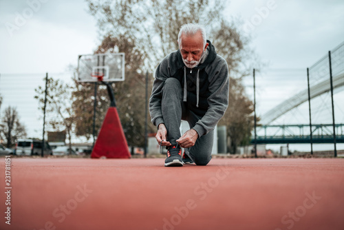 Active senior man in sportswear tying shoelaces before run outdoors.