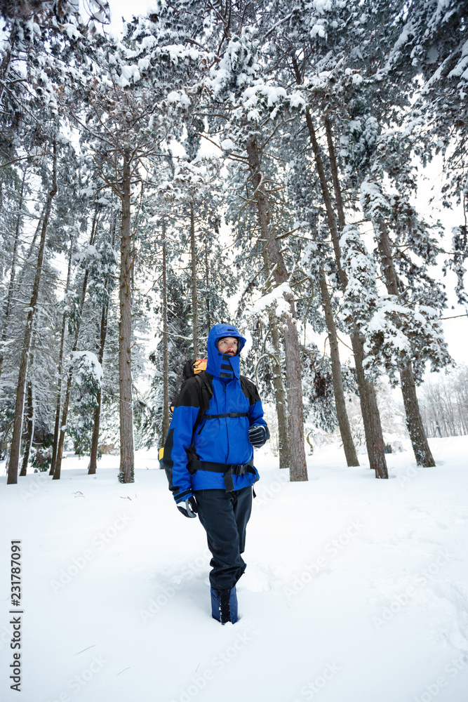 Young male mountain climber wearing blue and black clothing hiking towards the camera through deep powder snow in fir tree forest. Wide angle frontal view. Active lifestyle in cold weather.