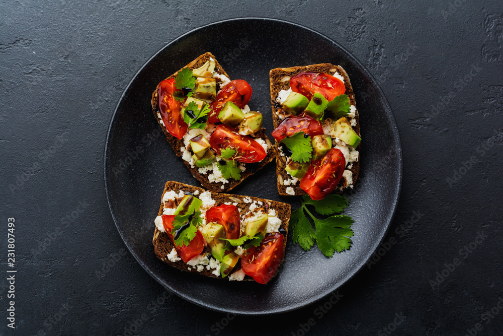 Avocados, cherry tomatoes and feta cheese sandwiches with balsamic sauce and cilantro on old olive board on dark black background. Top view. Avocados, cherry tomatoes and feta cheese sandwiches with balsamic sauce and cilantro on old olive board on dark black background. Top view.
