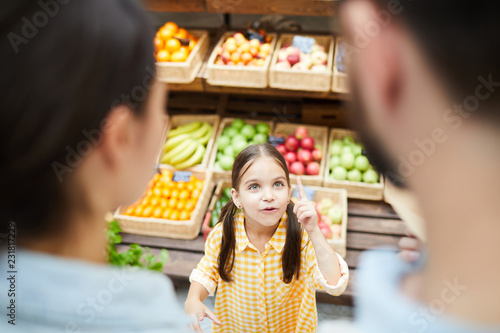 Cheerful excited girl with pony tails showing index finger while having idea in food store and offering it to parents