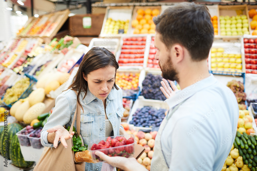 Puzzled frowning young female customer with shopping bag on shoulder displeased with quality of products and looking at strawberry shown by grocer in farmers market