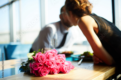 Bunch of pink roses on sunlit table and young amorous couple leaning over table for kiss photo