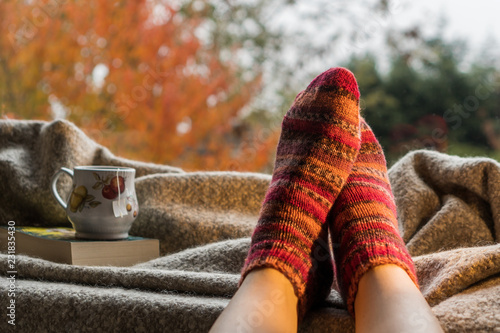 Woman's feet wearing handmade knitted colorful wool socks next to a hot cup of tea in a cozy decor. Keeping warm on a cold day concept. 