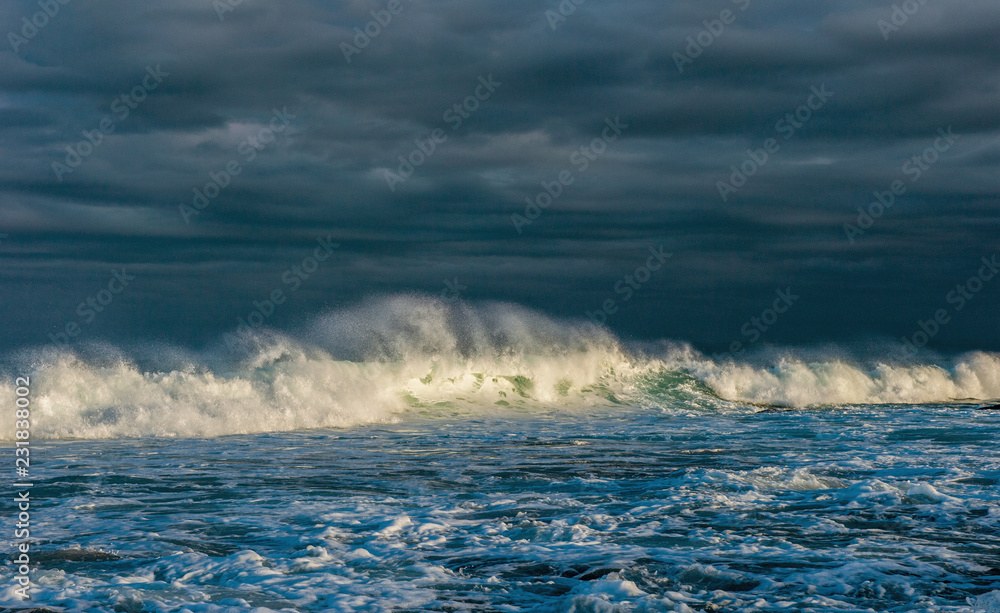Powerful ocean wave on the surface of the ocean. Wave breaks on a shallow bank. Stormy weather, clouds sky background. Seascape.