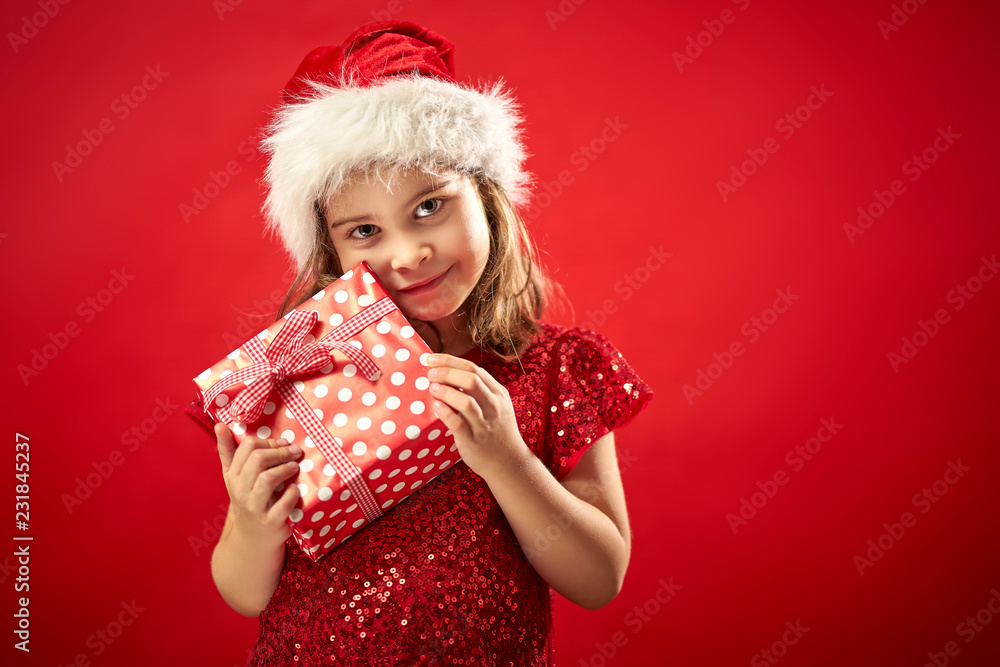 adorable little girl in a Christmas dress in a Santa's hat holding a present adorable little girl in a Christmas dress in a Santa's hat holding a present