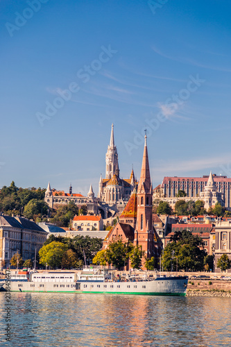 Two large Catholic churches on the Danube River in Budapest. The ship sails on the river. Many green plants on the Buda hill.