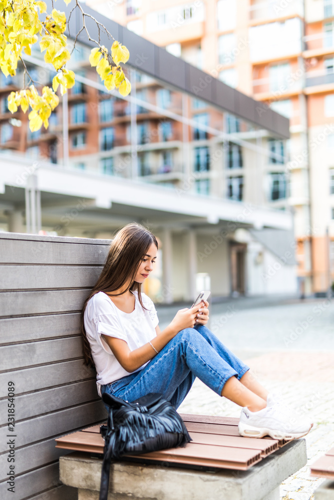 Beautiful young girl texting on the smartphone sitting in a bench in urban.