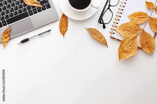 White desk copy space seen from above with a laptop keyboard, notebook, glasses, coffee, pen and yellow autumn leaves