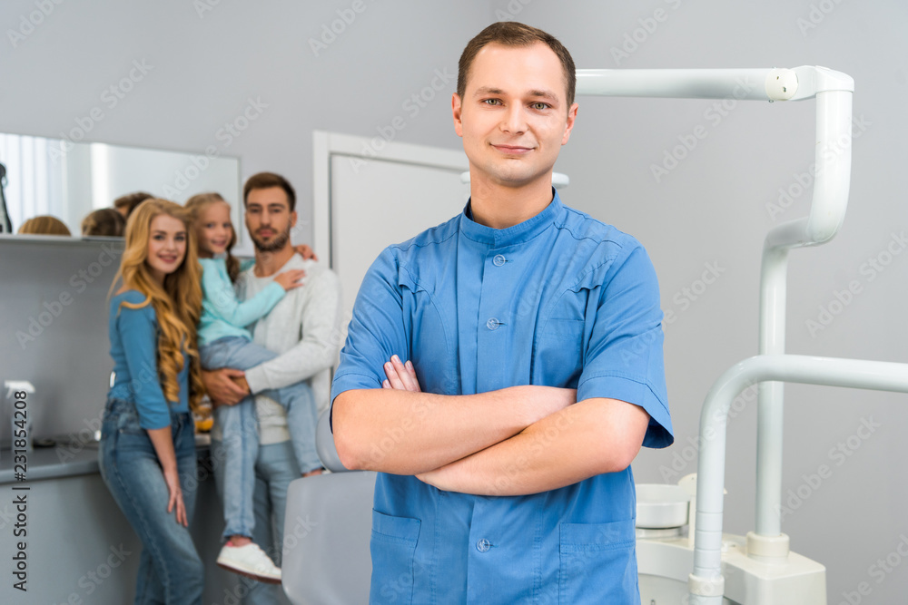 Fototapeta premium handsome young dentist with crossed arms looking at camera while young family standing on background together