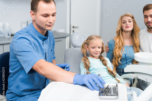 handsome young dentist with tools working with little child while her parents sitting near her