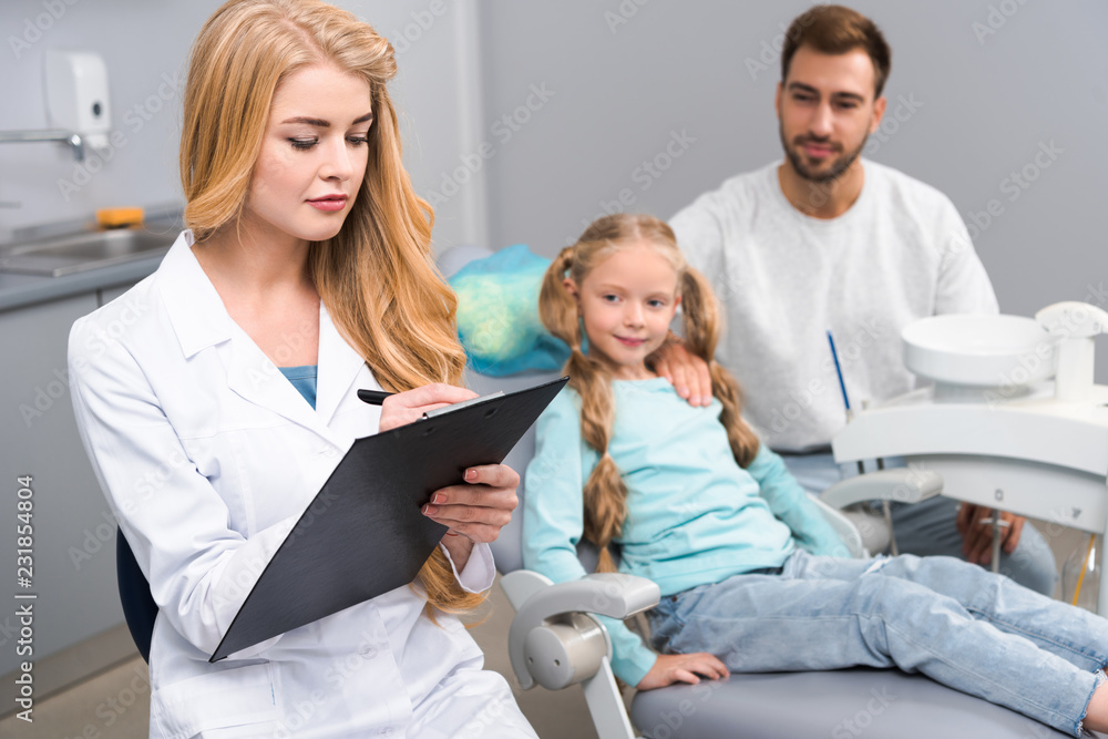 attractive female dentist writing in clipboard while little child and her father sitting on background
