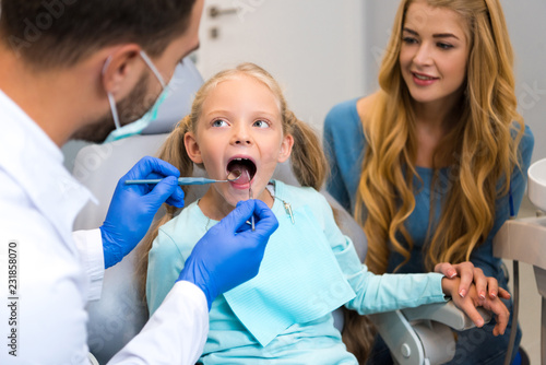 dentist examining teeth of little child while mother sitting near her