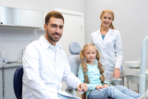 young male and female dentists with little child looking at camera in dental office