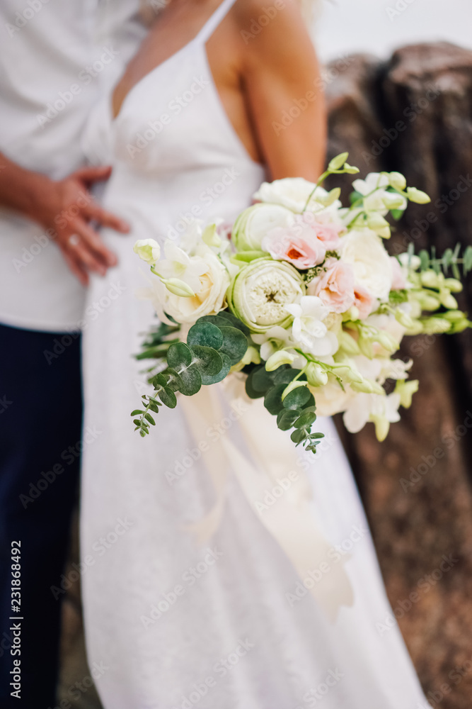 Wedding bouquet of white and pink flowers and greenery in the hands of the bride. In the background are a couple, the bride and groom Wedding bouquet of white and pink flowers and greenery in the hands of the bride. In the background are a couple, the bride and groom