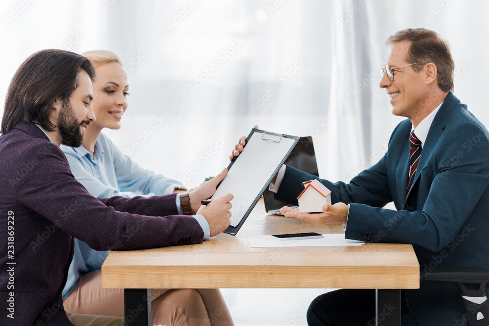 adult smiling insurance agent showing contract and small house to young couple in office adult smiling insurance agent showing contract and small house to young couple in office
