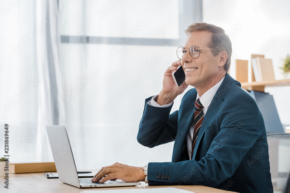 adult smiling businessman in glasses talking on smartphone and using laptop in office adult smiling businessman in glasses talking on smartphone and using laptop in office