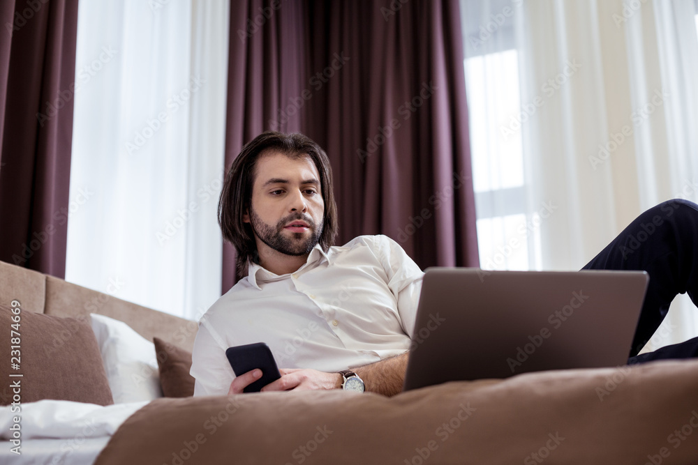 Successful business. Serious nice businessman looking at the laptop screen while lying on the bed in the hotel Successful business. Serious nice businessman looking at the laptop screen while lying on the bed in the hotel