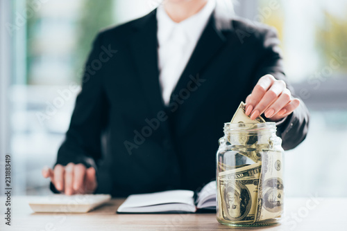 cropped shot of businesswoman working and putting dollar banknote in glass jar
