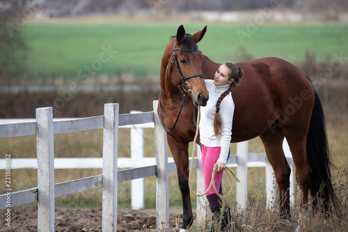 Young girl standing with her bay horse near paddock. Equestrian life concept background