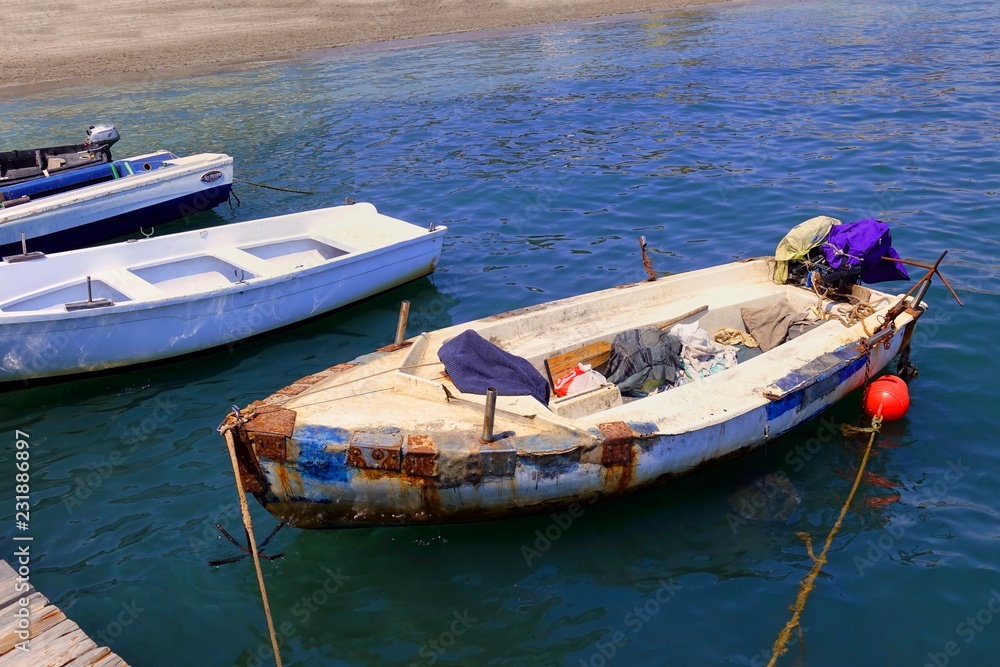Small boats moored at a wooden jetty on a bright blue sea - one of them is very old and patched with rusty metal plates