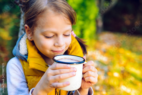 child girl drinking chocolate from a cup dressed in a warm yellow vest in autumn scenery