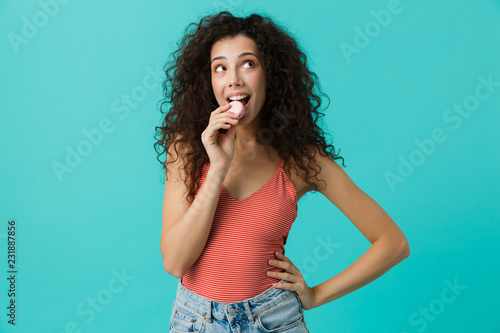 Photo of pretty woman 20s with curly hair smiling and eating macaron biscuit, isolated over blue background
