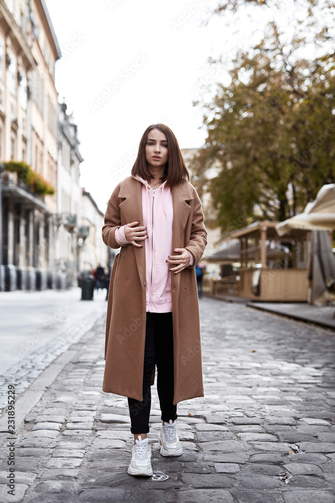 Portrait of a young woman in brown unbuttoned coat and pink hoody. Woman posing outdoors on the street at overcast autumn day