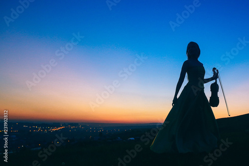 The girl in an evening dress holds a violin and bow in one hand, sunset, silhouette