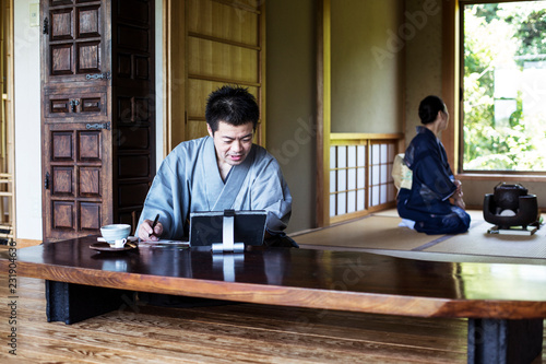 Japanese man wearing kimono sitting on floor in traditional Japanese house, looking at digital tablet. photo