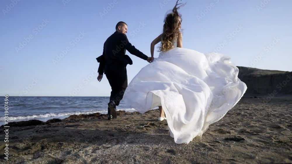 Following young couple in bridal dress and suit running on sandy beach, beautiful white dress waving on the wind Following young couple in bridal dress and suit running on sandy beach, beautiful white dress waving on the wind