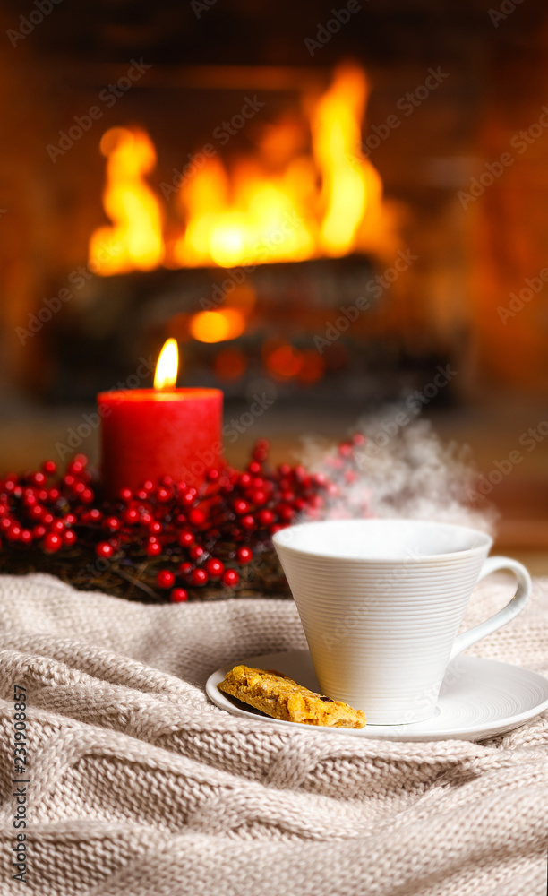 Cup of hot drink with steam and cookie berries red candle in Christmas decoration on knitted plaid in front of fireplace. Christmas New Year concept. Cozy relaxed magical atmosphere home interior. Cup of hot drink with steam and cookie berries red candle in Christmas decoration on knitted plaid in front of fireplace. Christmas New Year concept. Cozy relaxed magical atmosphere home interior.