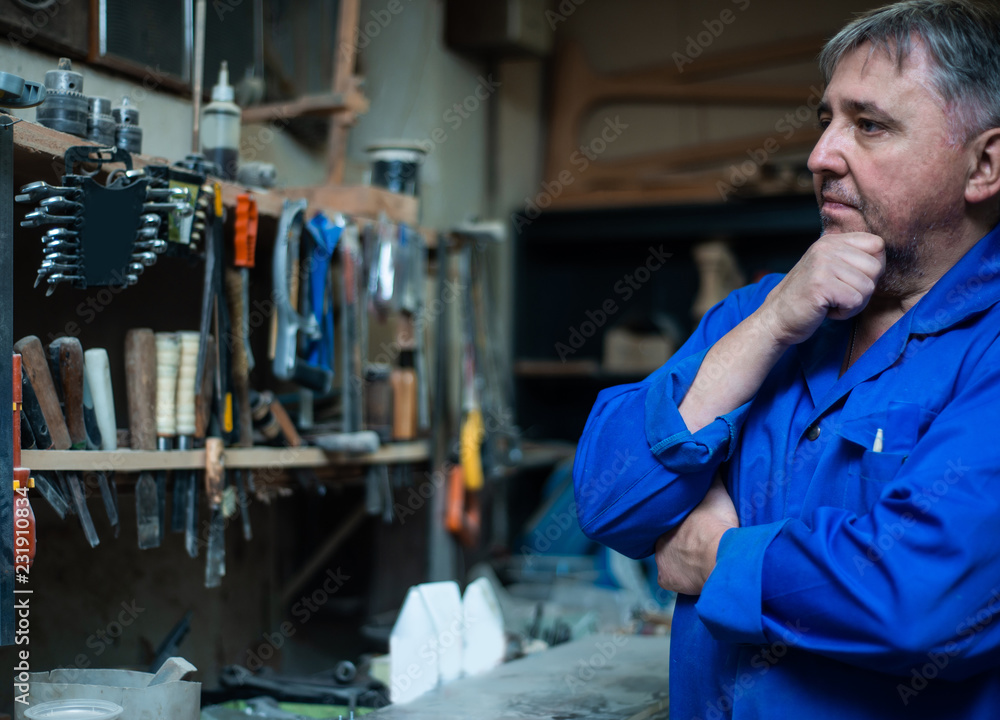 Mature man carpenter in overalls chooses tools in the workshop