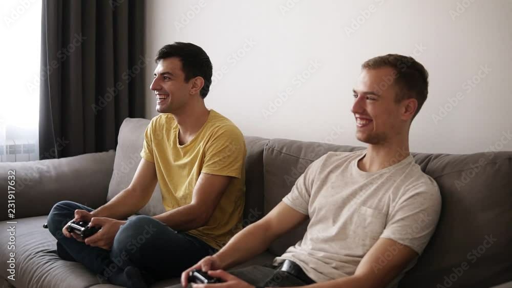 Two young guys playing video games at home, holding joysticks and sitting on the grey sofa in loft interior room.Smiling, excited about competition Two young guys playing video games at home, holding joysticks and sitting on the grey sofa in loft interior room.Smiling, excited about competition