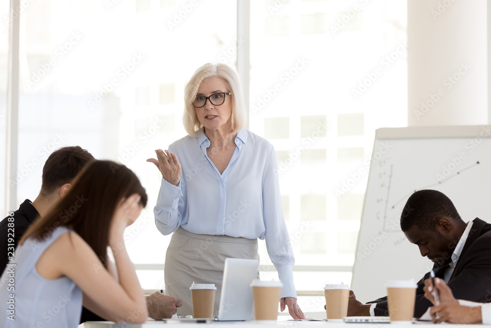 Angry mature businesswoman hold business briefing in office scolding young employees for bad work results, mad female boss lecture millennial workers or subordinates for failure at company meeting Angry mature businesswoman hold business briefing in office scolding young employees for bad work results, mad female boss lecture millennial workers or subordinates for failure at company meeting