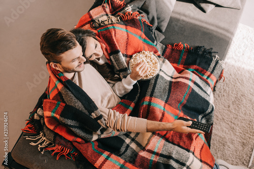 high angle view of beautiful young couple watching tv with popcorn on couch and covering with plaid