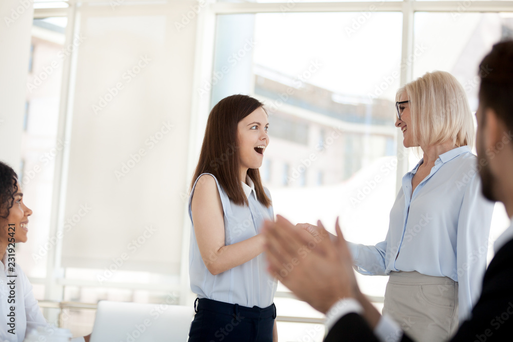 Fototapeta premium Excited millennial employee amazed by work promotion or personal achievement, smiling middle-aged businesswoman handshake happy young colleague congratulating with success. Reward concept