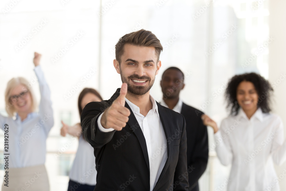 Excited Caucasian millennial employee or manager show thumbs up, supported by diverse colleagues cheering at back, happy candidate or applicant satisfied with interview, client pleased with service Excited Caucasian millennial employee or manager show thumbs up, supported by diverse colleagues cheering at back, happy candidate or applicant satisfied with interview, client pleased with service