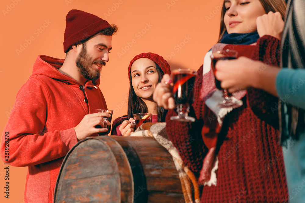 Fototapeta premium Smiling european men and women during party photoshoot. The guys posing as friends at studio fest with wineglasses with hot mulled wine on foreground.