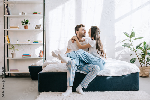 beautiful young couple in pajamas cuddling while sitting on bed in morning