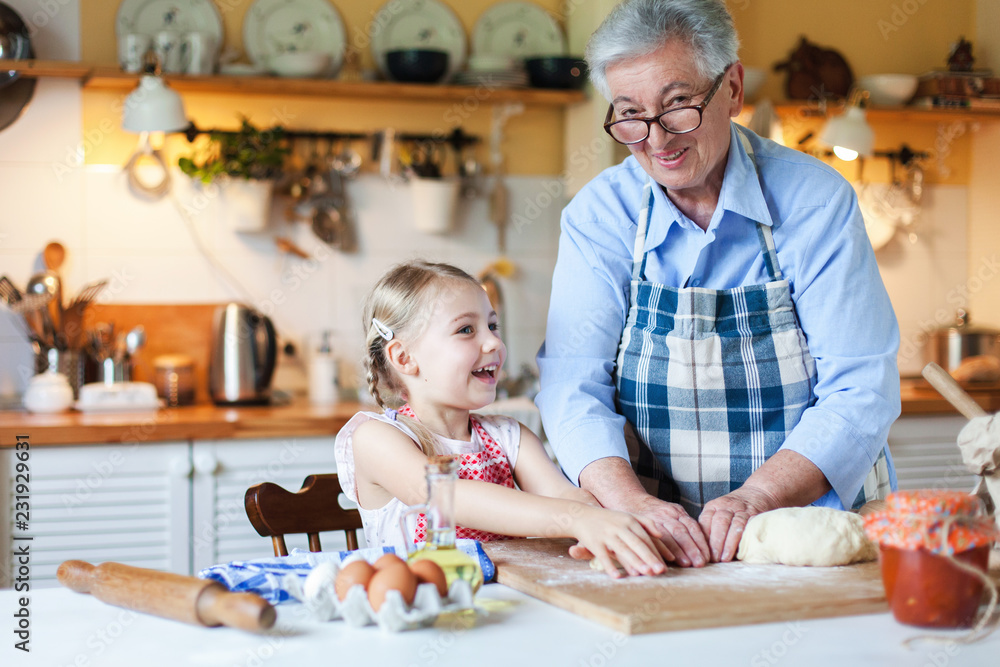 Grandmother and granddaughter are cooking in cozy home kitchen. Family is happy together. Senior woman and cute little child girl are smiling. Kid is helping to make bakery and homemade cakes. Grandmother and granddaughter are cooking in cozy home kitchen. Family is happy together. Senior woman and cute little child girl are smiling. Kid is helping to make bakery and homemade cakes.