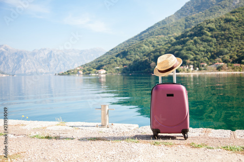 Fototapeta Naklejka Na Ścianę i Meble -  Pink suitcase with straw hat on sea beach. Concept of travel, vacation, female tourism, trip, journey, adventure. Nature background of amazing view with blue lake, coast, green mountains.